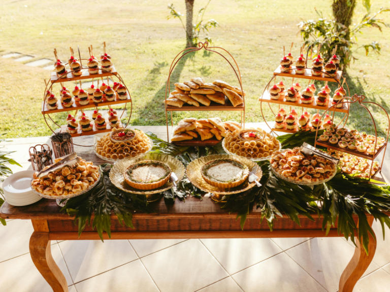 Buffet table with assorted pastries, mini burgers, tarts, and finger foods arranged on wooden and metal stands outdoors.