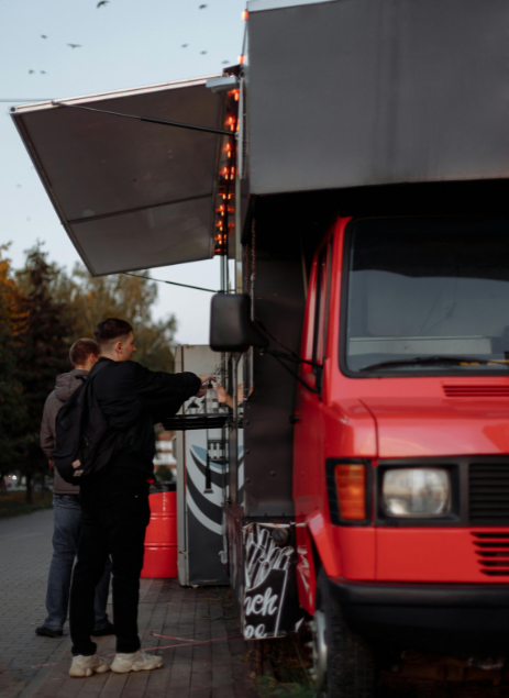 Two men order food from a red and black food truck parked on a sidewalk during early evening.