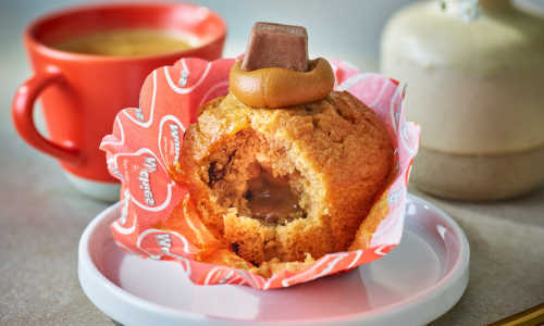 A partially eaten caramel-filled cupcake topped with caramel and chocolate, with a red mug and a white jar in the background.